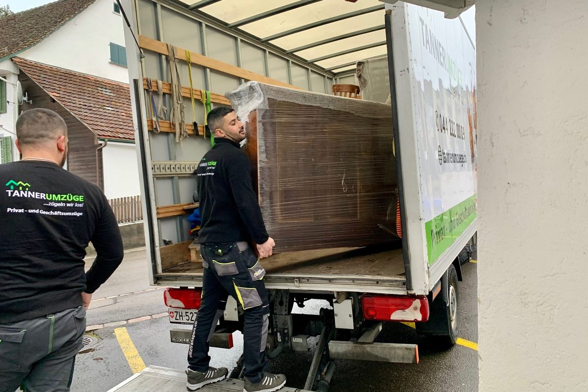 Zwei Mitarbeiter von Tanner Umzüge laden einen Schrank in den beschrifteten Umzugswagen