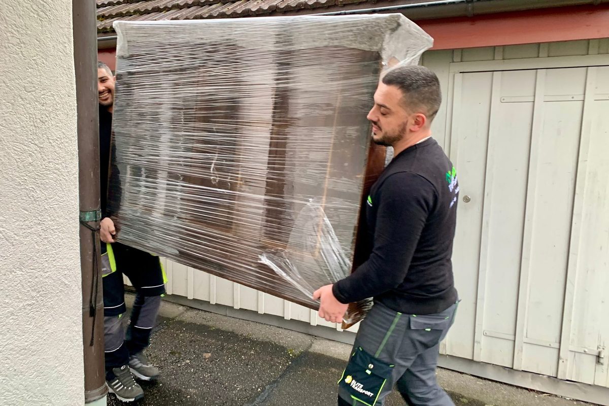 Transporte Zürich: Zwei Mitarbeiter von Tanner Umzüge tragen ein in Folie verpacktes Möbelstück zum Fahrzeug
