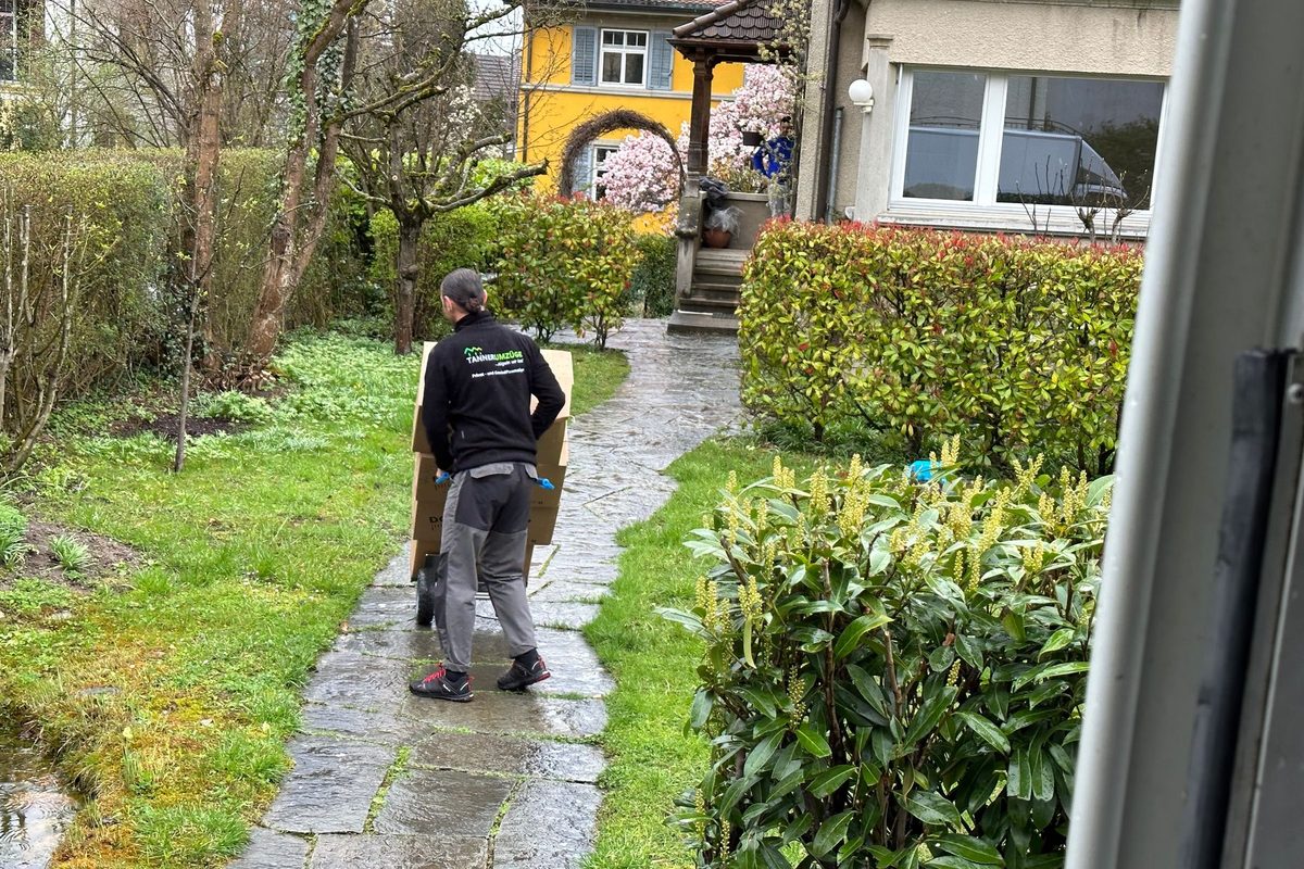 Umzugsfirma Zürichsee: Zwei Mitarbeiter von Tanner Umzüge tragen Umzugskartons über einen Gartenweg