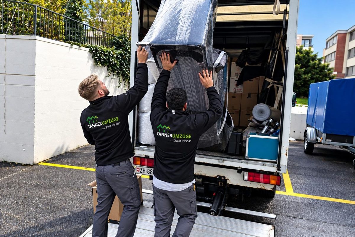 Zwei Mitarbeiter von Tanner Umzüge heben ein eingepacktes Möbelstück gemeinsam in den Wagen